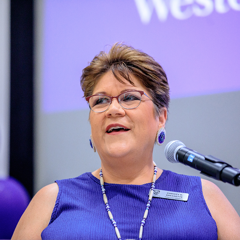 Person speaking into a microphone at a podium or table, with a purple presentation screen in the background displaying partial text and a balloon visible to the side.